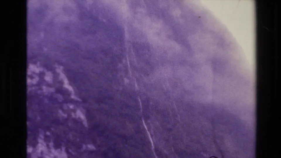 New Zealand aerial home-movie frame showing a cloudy mountain face, faint watercourse, and aged 8mm grain with purple color shift.