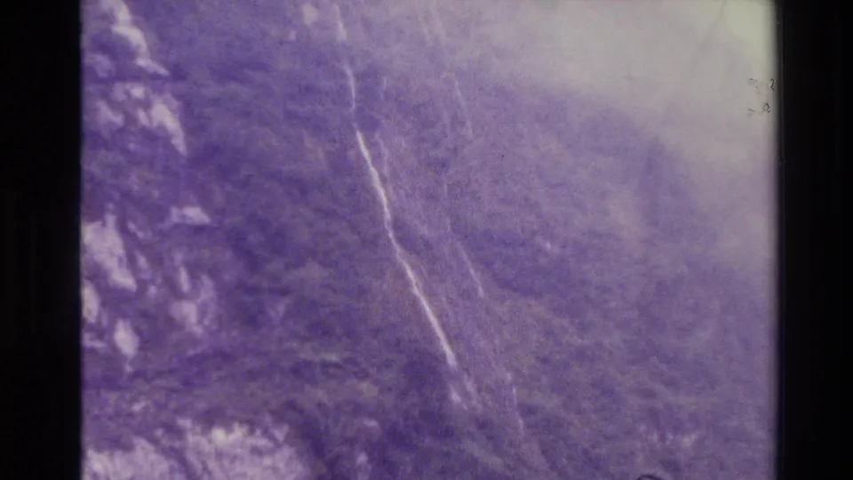 Aerial New Zealand 1982 landscape frame with mist, forested mountain contours, pale water traces, and visible aged film grain.