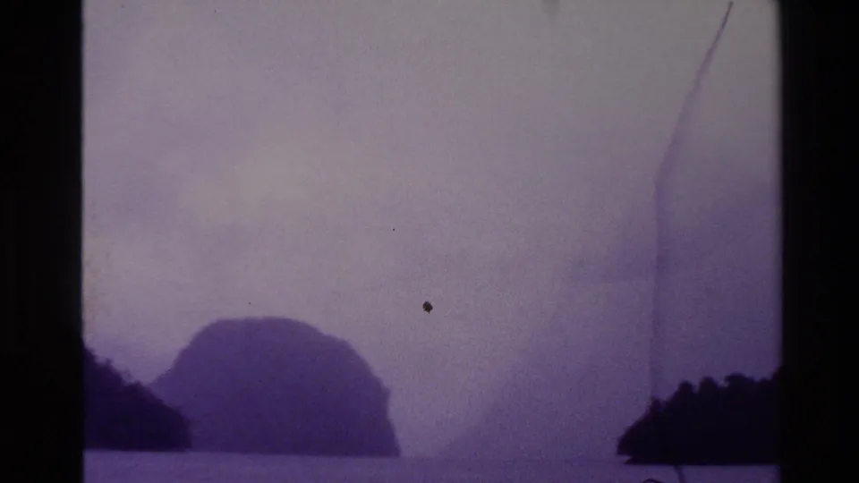 A faded 8mm view of New Zealand coastal islands partly obscured by mist, with dust specks and frame-edge artifacts visible.