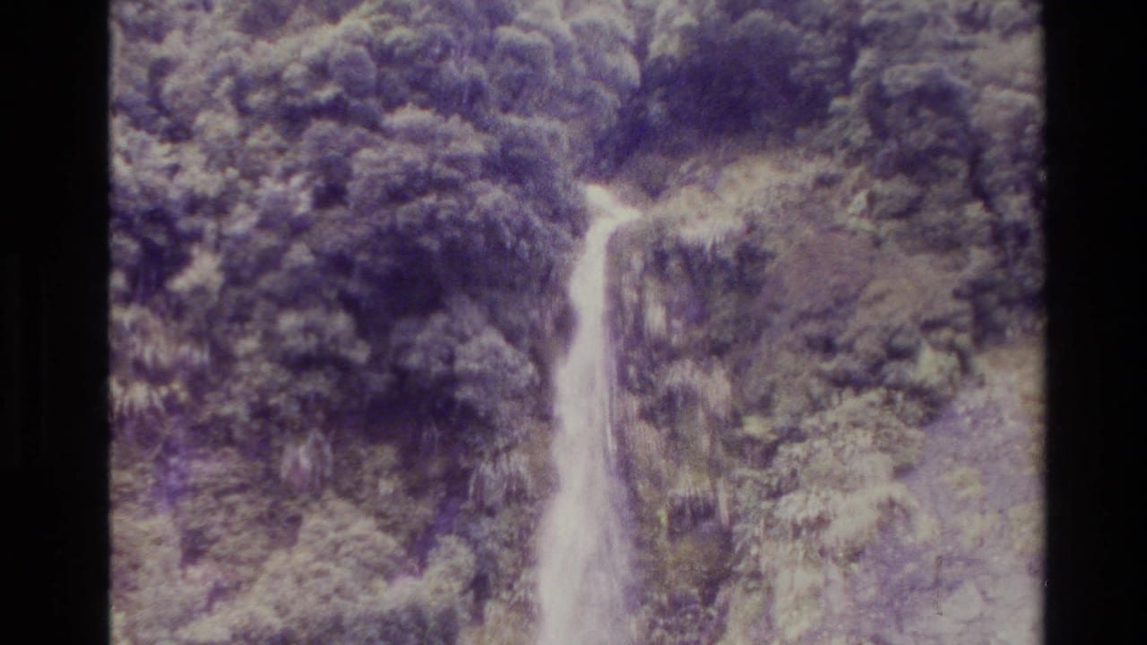 1982 New Zealand 8mm frame showing a long waterfall dropping through dense trees and rock, with faded purple-green film color.