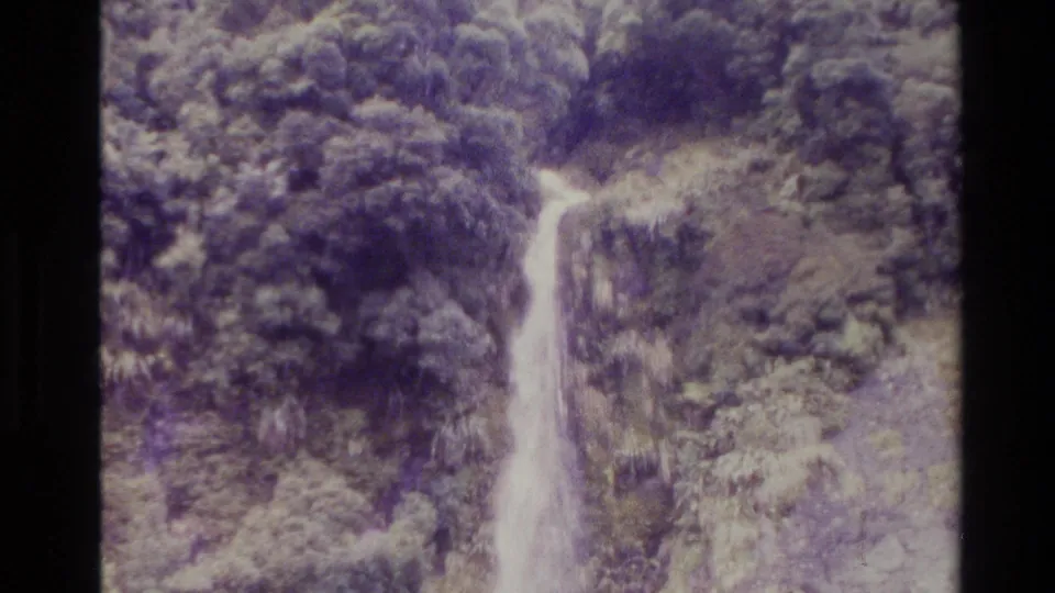 1982 New Zealand 8mm frame showing a long waterfall dropping through dense trees and rock, with faded purple-green film color.