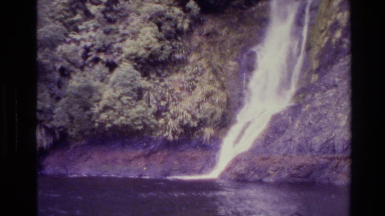 1982 New Zealand 8mm frame showing a waterfall cascading into a calm dark pool beneath dense forest growth.
