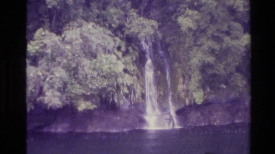 1982 New Zealand 8mm frame with a narrow waterfall, rocky bank, and thick vegetation above a dark river or pool.