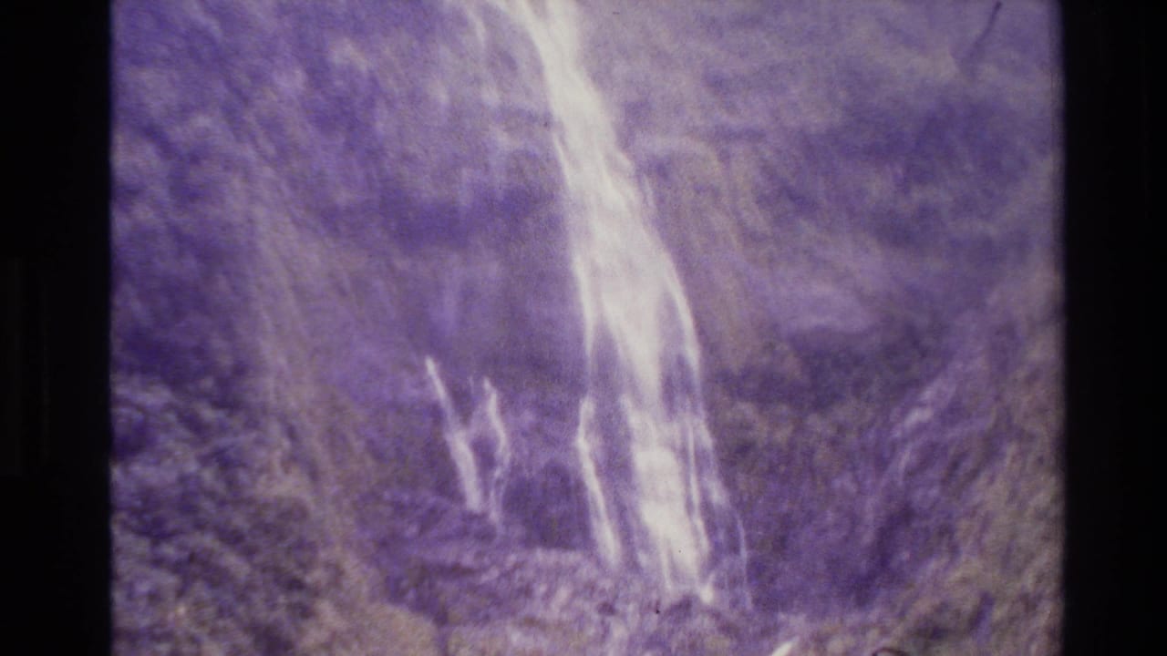 1982 New Zealand home-movie frame showing a pale waterfall on a steep, purple-shifted mountain slope.