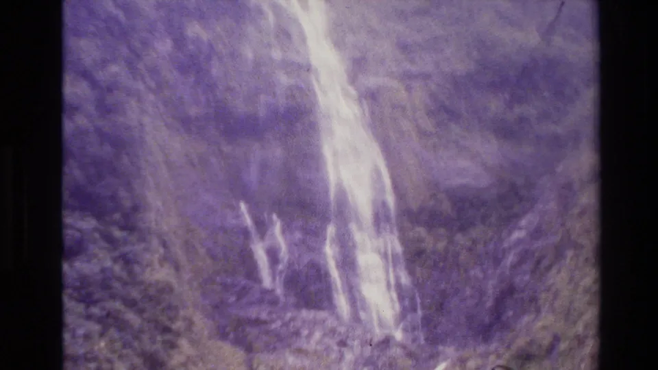 1982 New Zealand home-movie frame showing a pale waterfall on a steep, purple-shifted mountain slope.