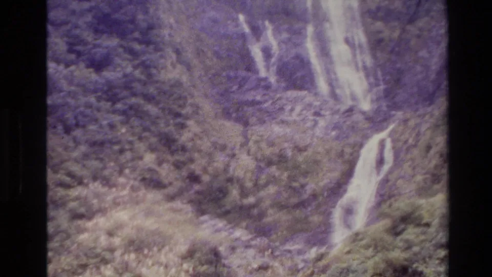 Home-movie view of multiple waterfall streams across a lush New Zealand hillside, with soft grain and color shift.
