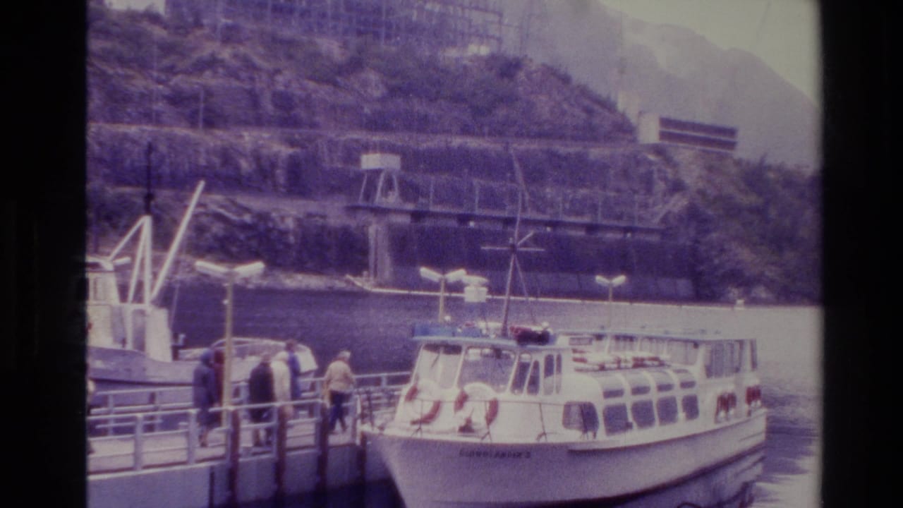 1982 New Zealand home-movie frame showing passengers on a dock near a moored white tour boat below steep mountain slopes.
