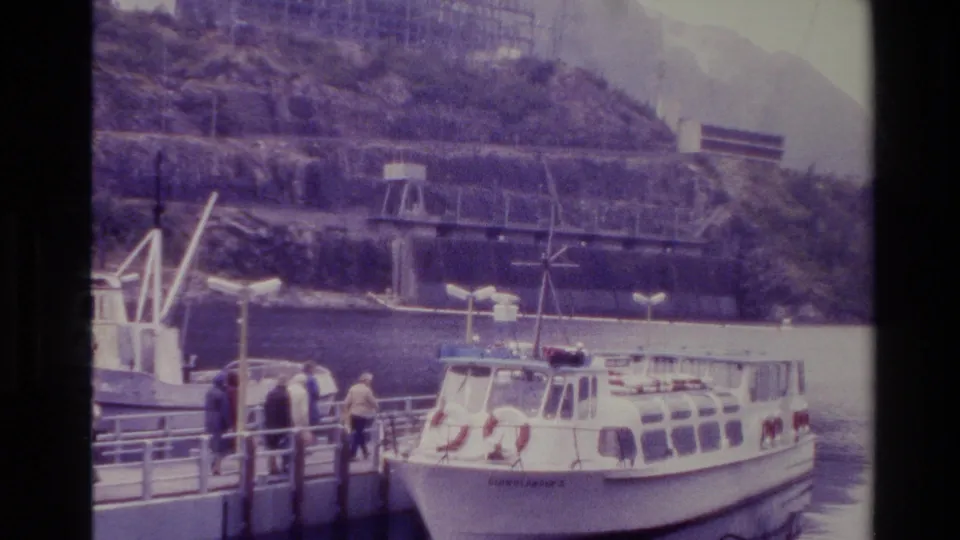 1982 New Zealand home-movie frame showing passengers on a dock near a moored white tour boat below steep mountain slopes.