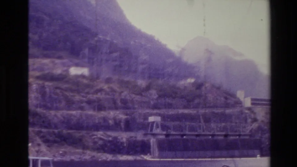 1982 New Zealand frame looking across calm water toward steep mountains, a small pier structure, and construction on the hillside.