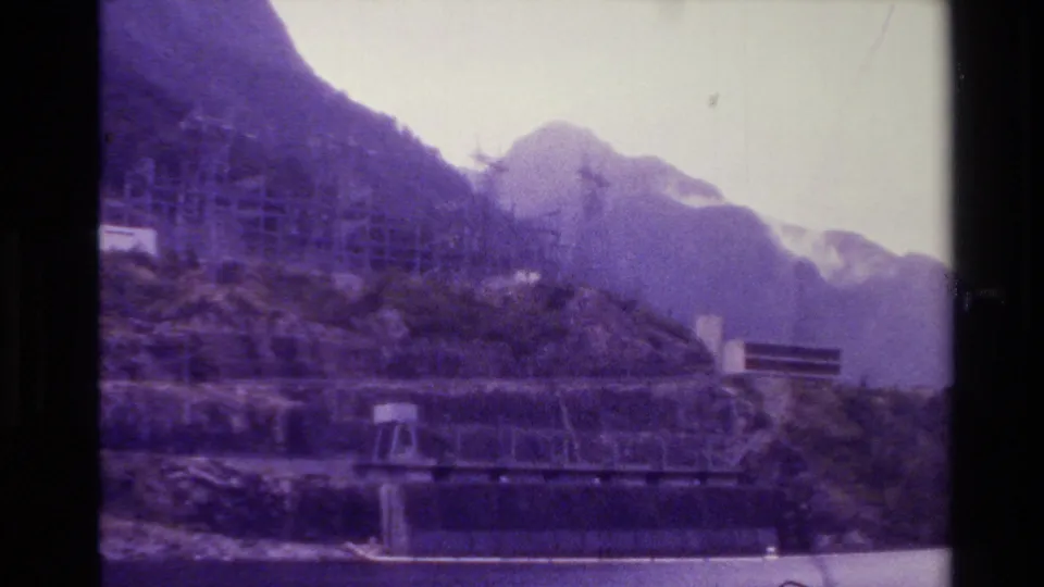 1982 New Zealand home-movie view of a steep mountain shoreline with construction scaffolding or framework above the water.