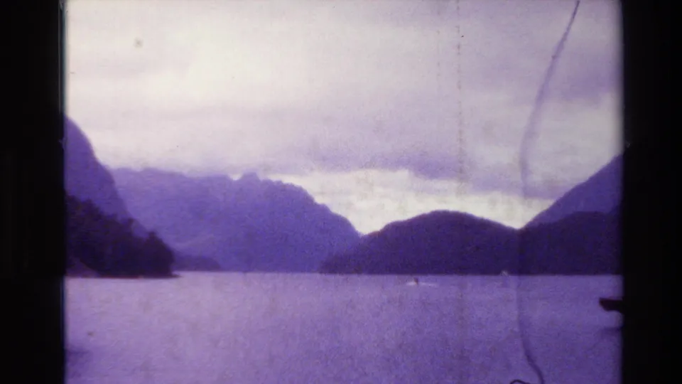Wide 1982 New Zealand home-movie view across calm water toward layered mountain peaks, with a small vessel in the distance.