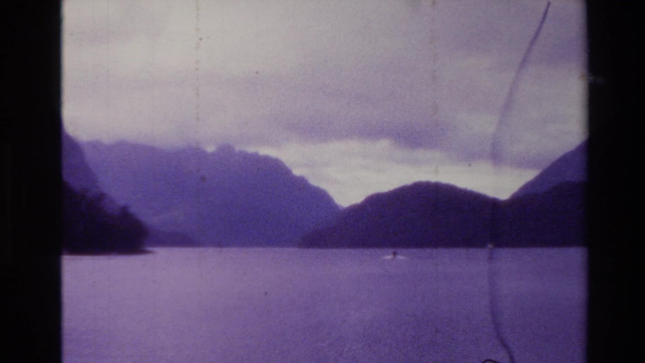 1982 New Zealand 8mm frame showing calm coastal water, dark mountain silhouettes, cloudy sky, and visible film scratches.
