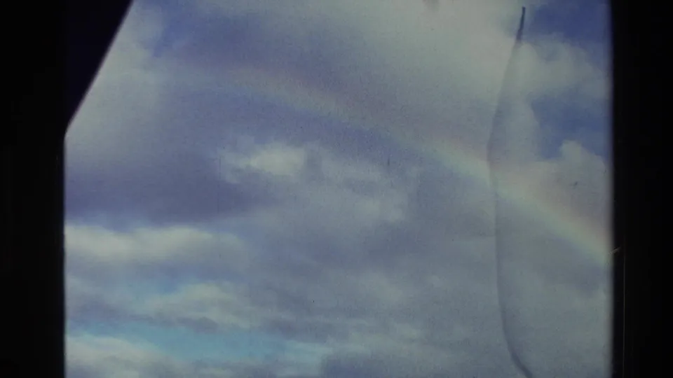 1982 New Zealand 8mm frame tilting upward to a faint rainbow across layered clouds, with dark frame edge and film wear visible.