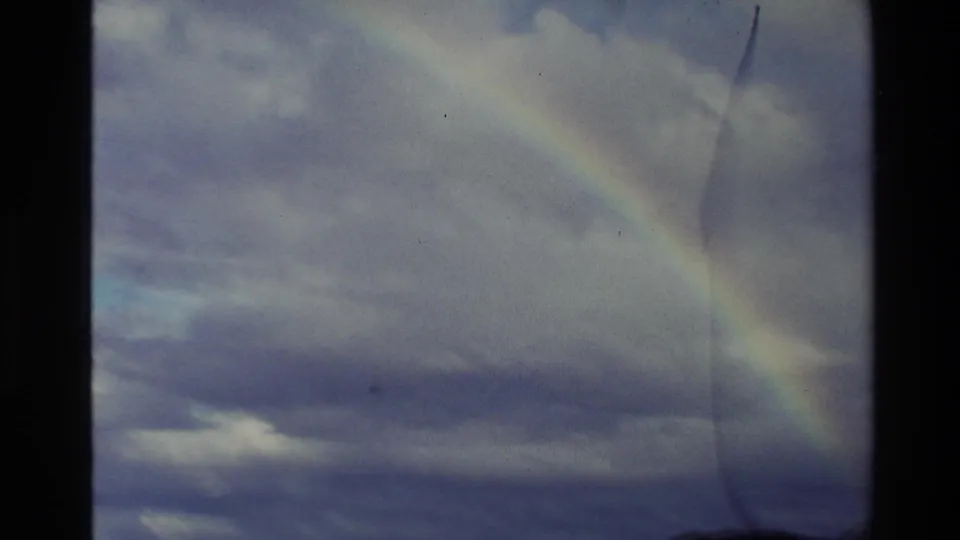 1982 New Zealand 8mm frame showing a brighter rainbow segment against heavy clouds, with grain, fading, and vertical scratches.
