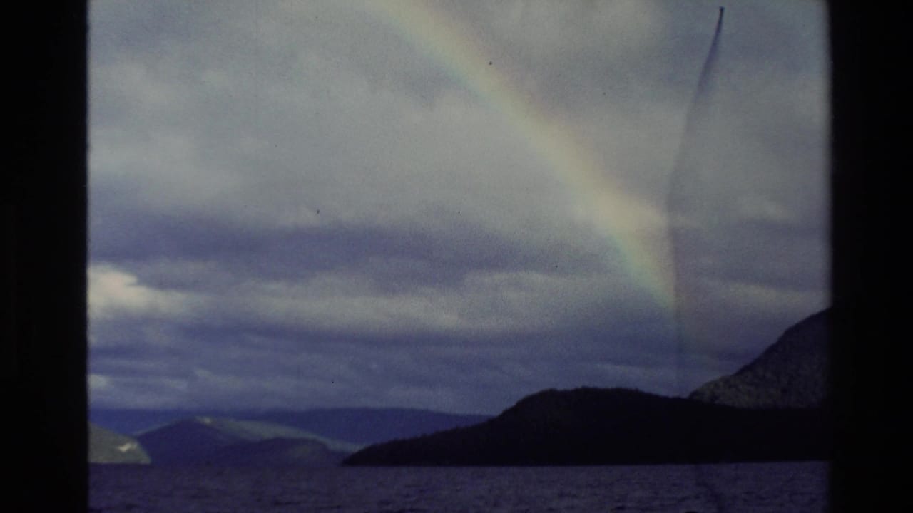 1982 New Zealand 8mm frame showing a faint rainbow arcing above a lake, silhouetted hills, and distant mountains under heavy cloud.