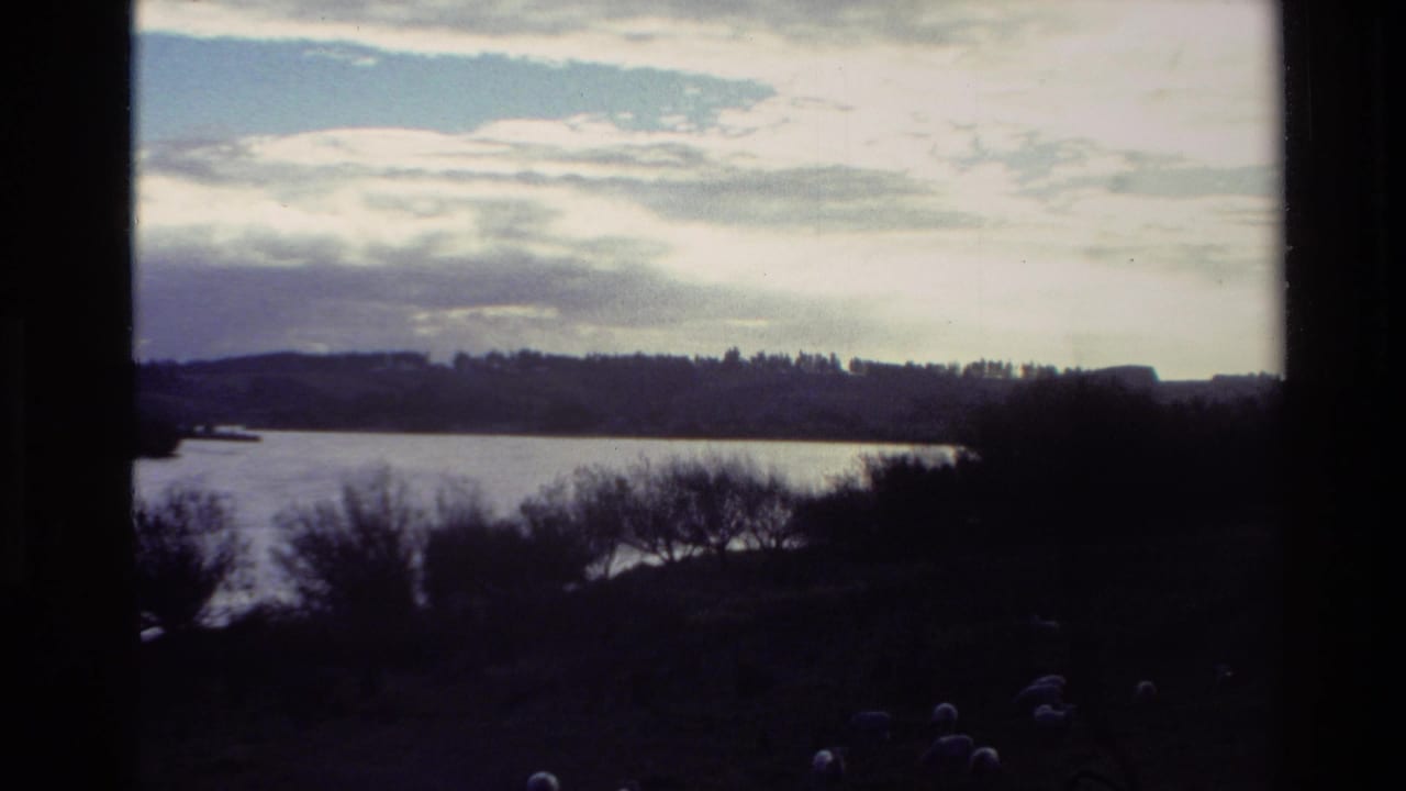 Wide river landscape with silhouetted trees, distant hills, cloudy sky, and dark film borders.