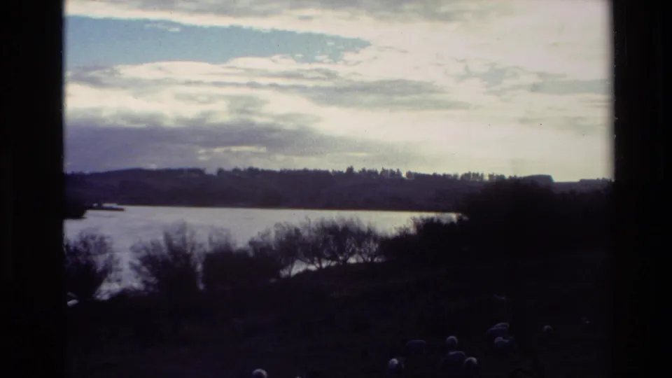 1982 New Zealand 8mm frame showing a broad river, dark shoreline trees, low hills, cloudy sky, and heavy gate borders.