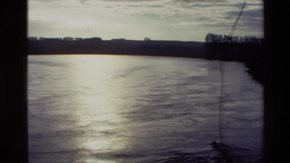 1982 New Zealand river view with bright reflected light on the water, a dark far shoreline, low hills, and visible film wear.