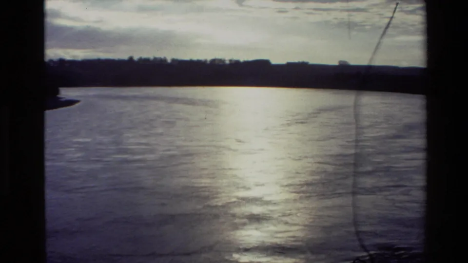 1982 New Zealand 8mm frame of a broad river bend with shimmering water, dark distant bank, cloudy sky, and gate-edge borders.