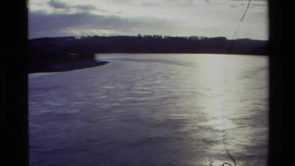 1982 New Zealand river scene showing a gentle curve, reflective water, silhouetted banks, distant trees, and visible 8mm scratches.