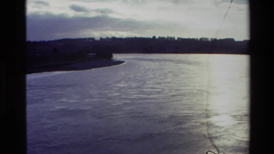 1982 New Zealand 8mm frame showing a broad waterway with shoreline and cloudy sky, bordered by dark film edges.