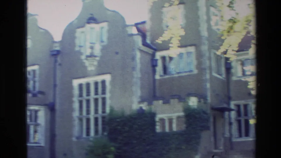 Soft 8mm frame of Olveston's exterior in Dunedin, showing tall windows, gabled rooflines, shrubs, and partial tree cover.