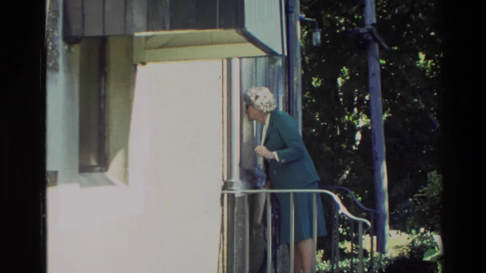 Adult woman in a green suit and white hat at a New Zealand residential doorway, framed by porch railings and exterior wall.
