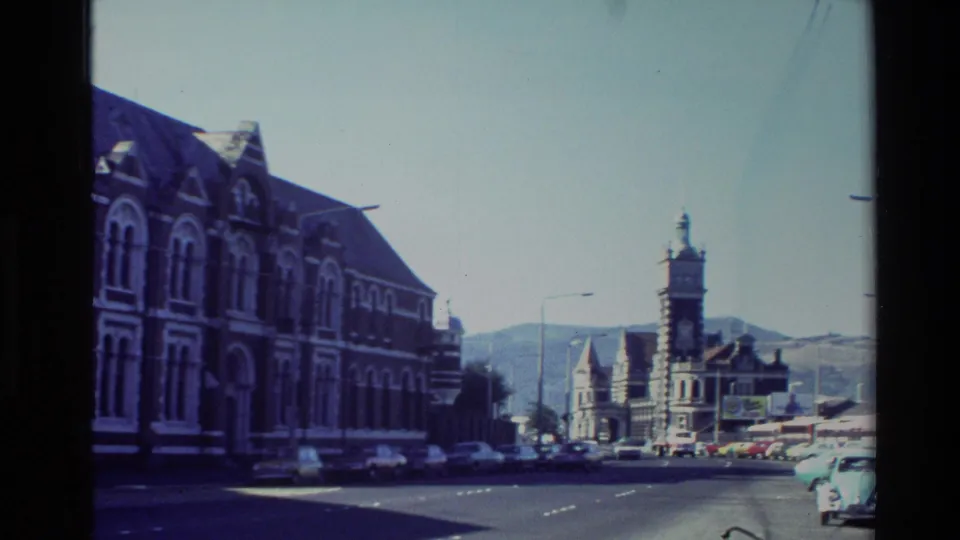 1982 New Zealand street view with older cars, ornate buildings, a prominent clock tower, streetlights, and a hillside in the distance.