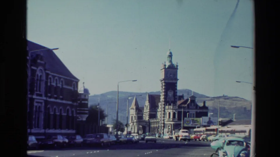 New Zealand 8mm street frame showing parked and moving cars near ornate town buildings, a clock tower, streetlights, and hills beyond.