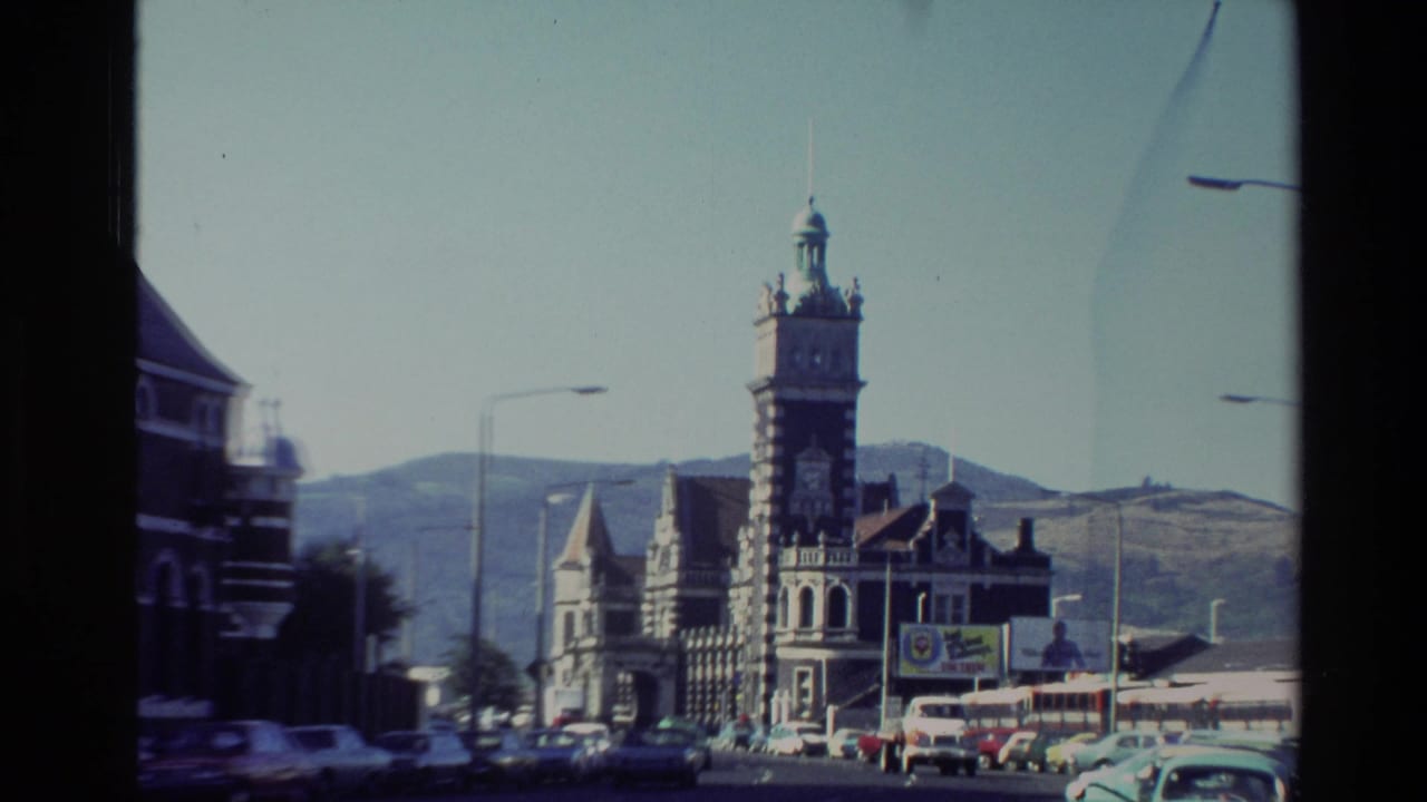 1982 New Zealand 8mm frame showing an ornate grey stone landmark with a clock tower, street traffic, signs, and hills in the background.