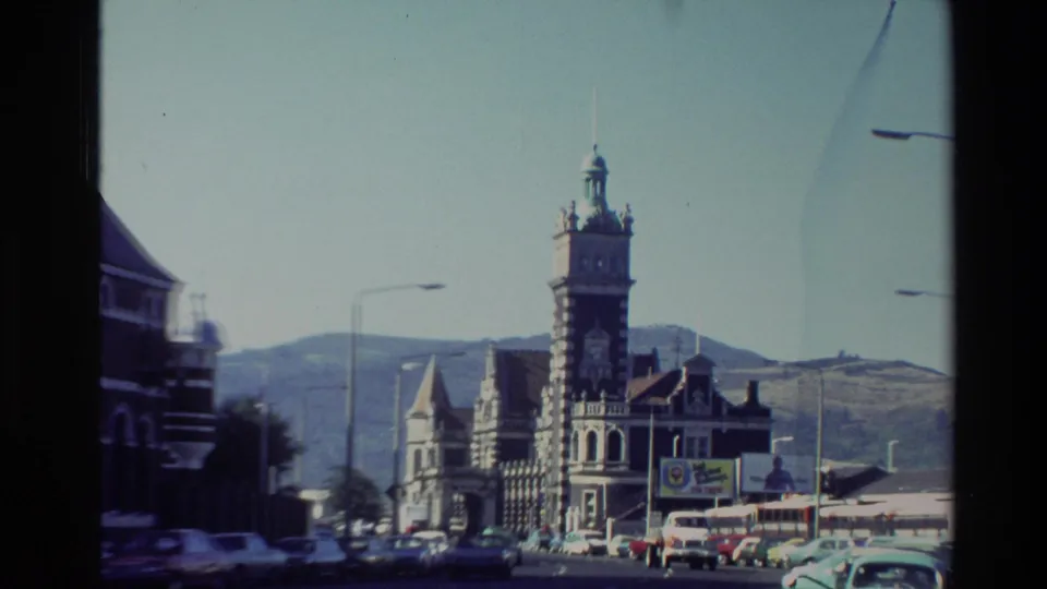 1982 New Zealand 8mm frame showing an ornate grey stone landmark with a clock tower, street traffic, signs, and hills in the background.