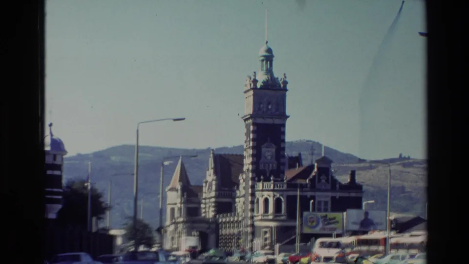1982 New Zealand street view of an ornate grey stone landmark with clock tower, visible road traffic, billboards, and hillside backdrop.