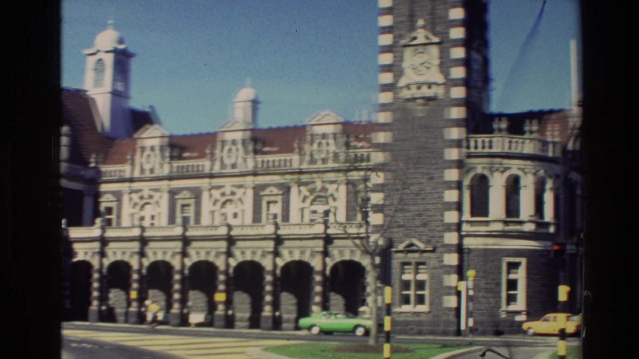 1982 New Zealand home-movie frame showing an ornate stone facade, clock tower, street crossing, and a bright green car below.