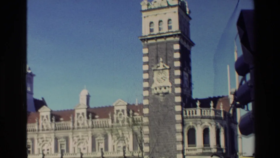 1982 New Zealand street view of a striped clock tower and decorative building facade with a yellow car passing below.