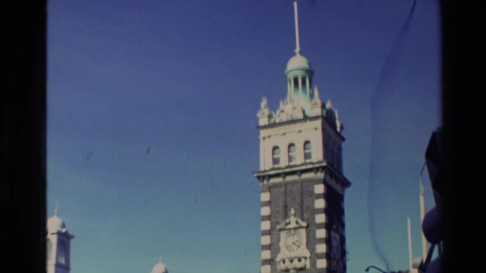 1982 New Zealand home-movie frame centered on the tall striped clock tower rising above the historic building.