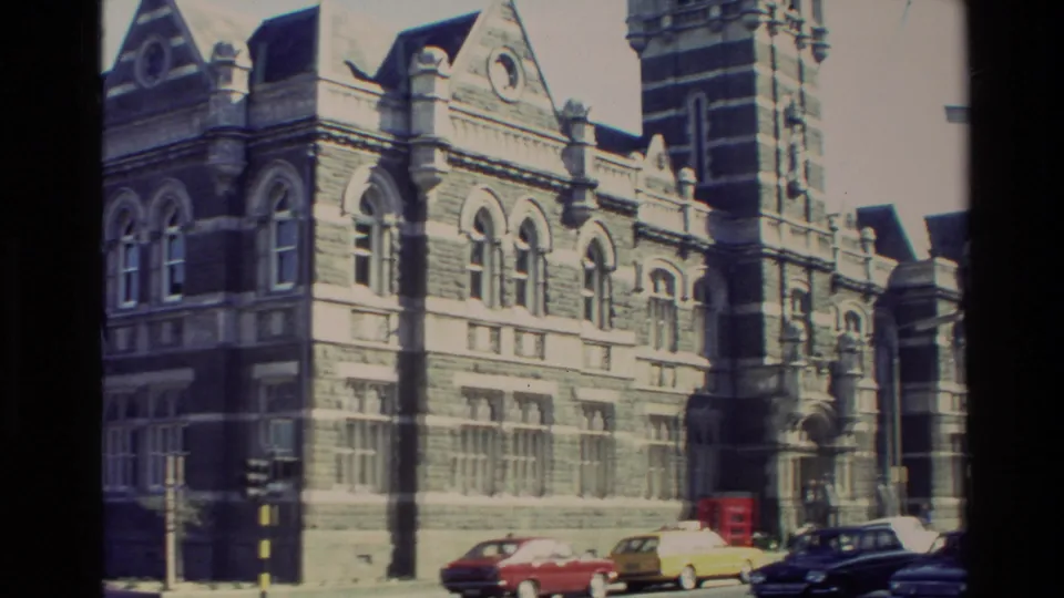 1982 New Zealand street-side view of the ornate historic building, arched windows, tower edge, and several period cars.