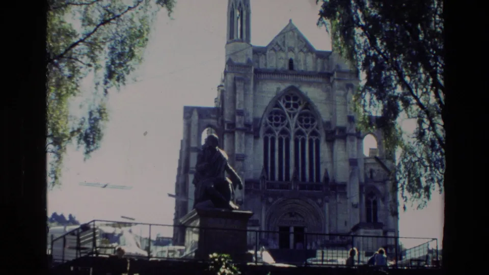 1982 New Zealand 8mm frame showing a Gothic-style church entrance behind a statue, with railings, trees, and small visitors near the base.