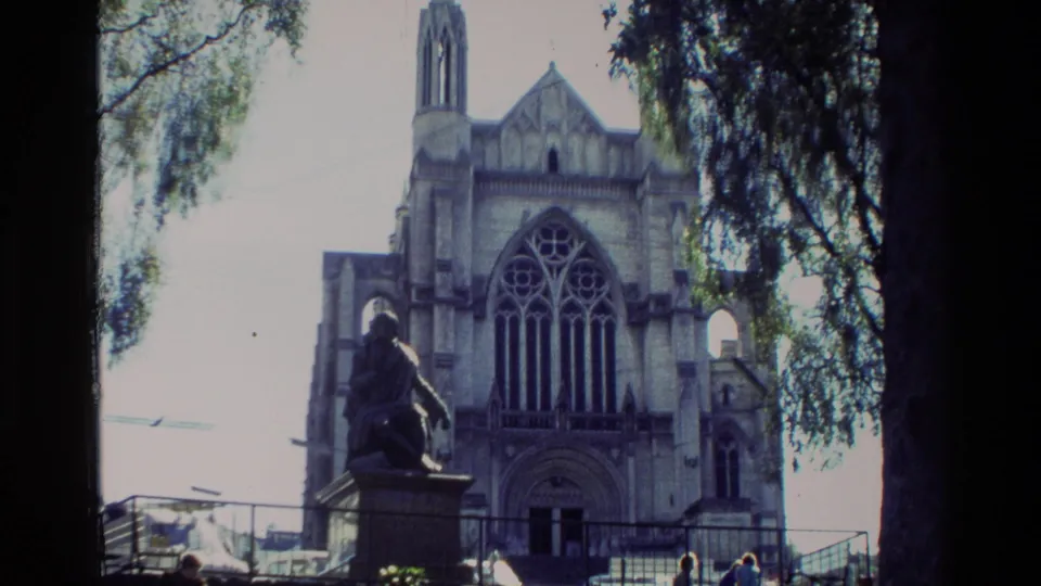1982 New Zealand 8mm frame of a Gothic-style church facade with a prominent foreground statue, leafy branches, railings, and distant visitors.