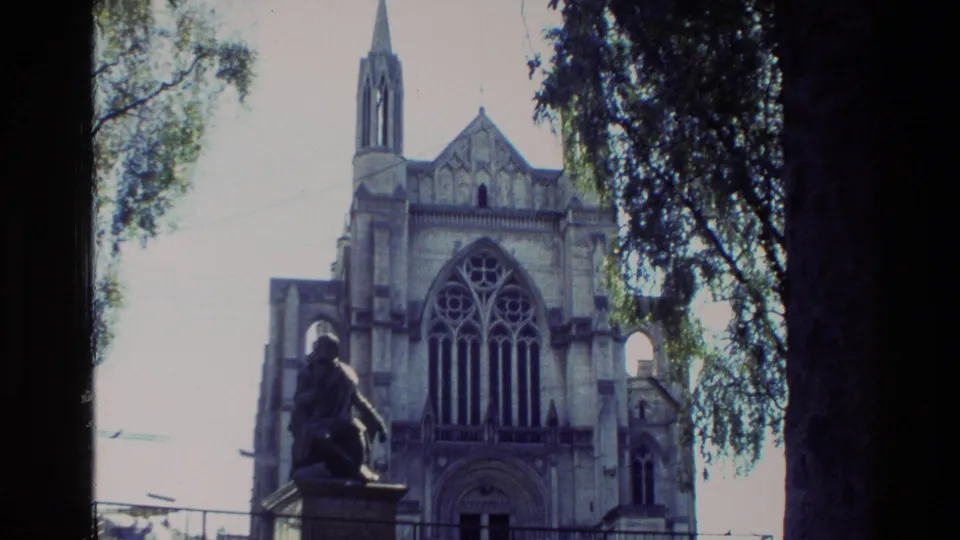 1982 New Zealand 8mm frame showing a Gothic-style church facade, arched window, spire, trees, and a foreground statue.