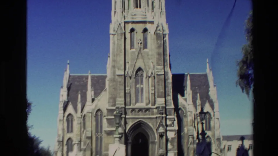 1982 New Zealand 8mm frame of a Gothic-style church tower, pointed entrance arch, roofline, lamps, and clear blue sky.