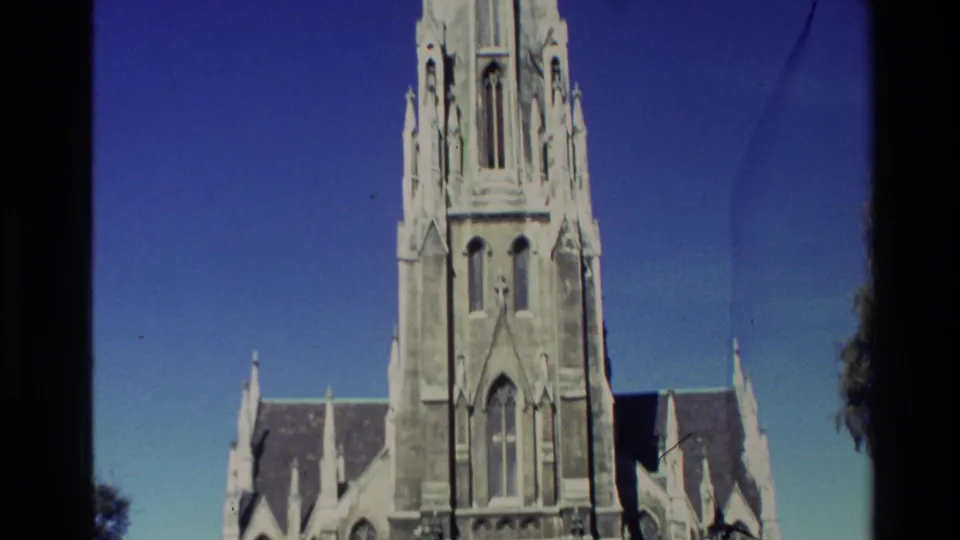 1982 New Zealand 8mm frame centered on the church tower, upper stonework, roof peaks, blue sky, and dark frame edge.