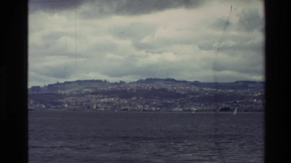 1982 New Zealand 8mm frame showing a distant lakeside town on hills, sailboats on the water, and an overcast sky.