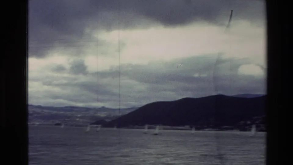 Small sailboats move across a cloudy New Zealand bay in 1982, with dark coastal hills along the far shore.
