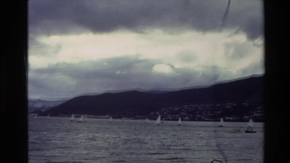 A home-movie view of sailboats on choppy New Zealand bay water, with a distant town at the base of coastal hills.