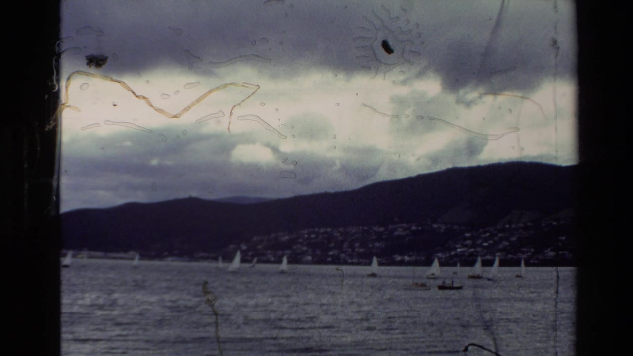 1982 New Zealand home-movie view of a bay with small sailboats, distant hills, shoreline buildings, cloudy sky, and visible film-edge wear.