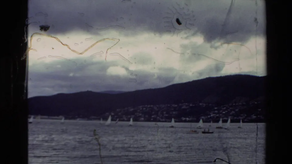 1982 New Zealand home-movie view of a bay with small sailboats, distant hills, shoreline buildings, cloudy sky, and visible film-edge wear.
