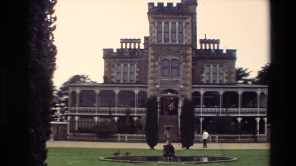 1982 New Zealand home-movie frame of a grand stone house with tower and verandas, framed by dark trees as a person walks across the grounds.