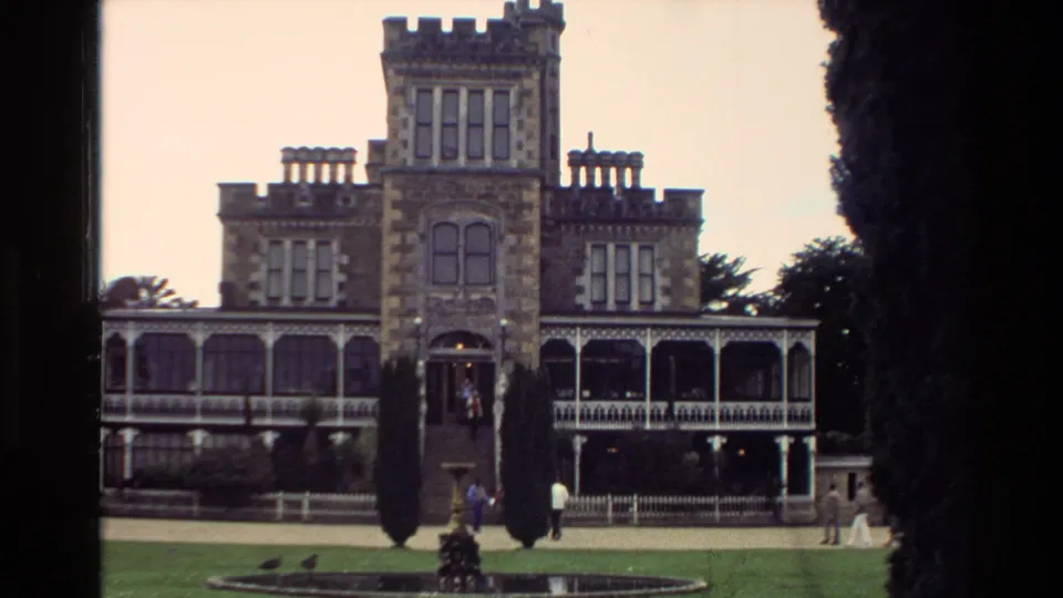 1982 New Zealand 8mm frame showing the mansion entrance and tower, with visitors near the steps and lawn in a casual travel view.