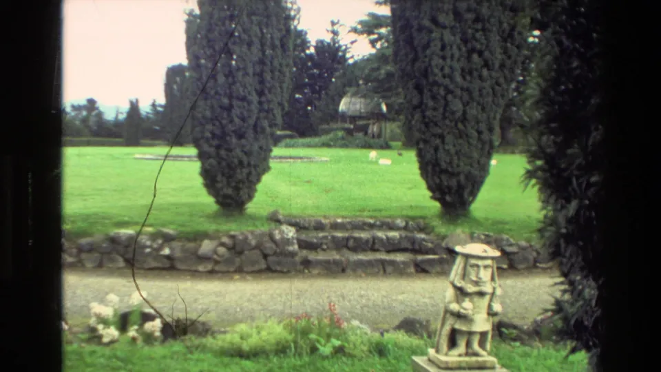1982 New Zealand 8mm frame of landscaped grounds near the mansion, with evergreens, grass, a gravel path, stone edging, and a small garden statue.