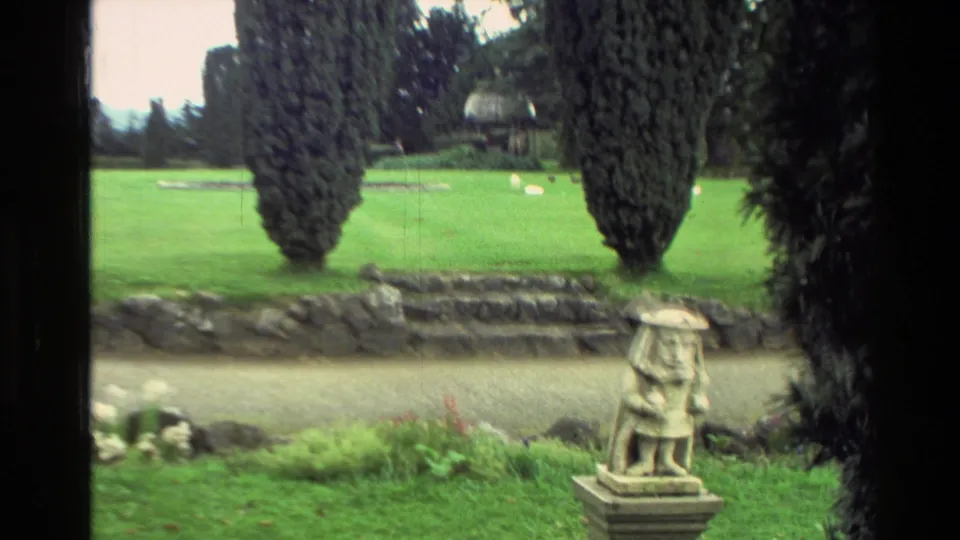 1982 New Zealand home-movie frame showing a small stone statue in landscaped grounds with rock steps, lawn, and tall trees.